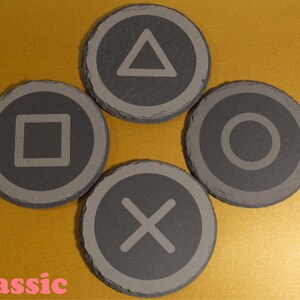 May include: Four grey slate coasters featuring etched PlayStation controller symbols: triangle, square, circle, and X. The coasters are arranged on a gold surface. The word "Classic" is written in pink.