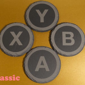 May include: Four circular slate coasters, each engraved with a letter: X, Y, B, and A. They are arranged in a cross formation against a gold backdrop. The word "Classic" is written in pink.