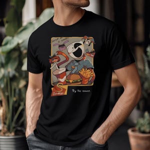 Op de afbeelding: Zwart T-shirt met een cartoon illustratie van een personage dat fastfood serveert. Het ontwerp bevat een hamburger, friet en een drankje, met de tekst "Serve with a smile!" en "Try the sauce." Het shirt is gemaakt van zacht materiaal.