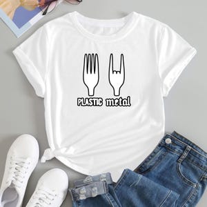 May include: White t-shirt featuring a black graphic of a fork and a hand making a rock and roll sign, with the text "PLASTIC metal" underneath. The shirt is styled with blue denim jeans and white trainers.
