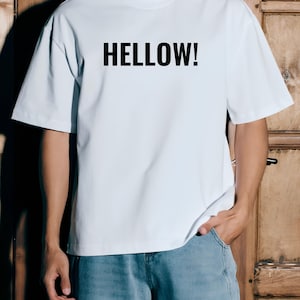 Puede incluir: Una camiseta blanca de manga corta con la palabra HELLOW! impresa en letras mayúsculas negras y negritas en el pecho. La prenda tiene cuello redondo y un corte holgado, combinada con unos vaqueros azules.