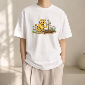May include: A white short-sleeve t-shirt featuring a graphic of a yellow bear wearing a polka-dot headband and green apron, gardening with a small shovel near a sapling, a watering can, and colorful flowers with a sign that reads GROW HOPE.