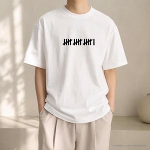 Può includere: Una t-shirt bianca a maniche corte con una stampa grafica nera sul petto. Il disegno è composto da tre gruppi di cinque segni di conteggio e un ulteriore segno singolo, per un totale di sedici linee verticali in fila orizzontale.