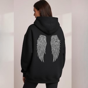 Könnte beinhalten: Schwarzer Kapuzenpullover mit einem großen, detaillierten silberfarbenen Flügeldesign auf dem Rücken. Die Flügel sind symmetrisch und haben ein gefiedertes Aussehen. Der Pullover hat eine Kapuze mit Kordelzug und eine lockere Passform.