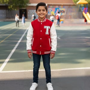 May include: A red and white letterman jacket with a large white "T" patch on the chest. The jacket has white sleeves, a white collar, and white stripes on the cuffs and hem. The jacket is worn with dark blue jeans and white sneakers.