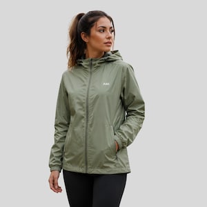 May include: An olive green lightweight hooded windbreaker jacket featuring a full-length front zip, elasticated cuffs, and two side pockets. The word AMI is printed in white on the upper left chest area of the garment, which is worn over black leggings.