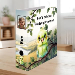 Può includere: Un raccoglitore personalizzato con un'illustrazione ad acquerello di un parrocchetto verde appollaiato su un ramo. Il raccoglitore include una foto di un bambino e il testo "Ben's schöne Kindergartenzeit". Il raccoglitore è su un tavolo di legno.