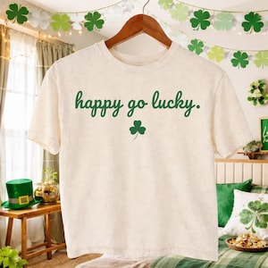 Può includere: T-shirt color crema con la scritta verde "happy go lucky" e un piccolo trifoglio verde. La maglietta è appesa a una gruccia di legno. Lo sfondo include decorazioni per la festa di San Patrizio.