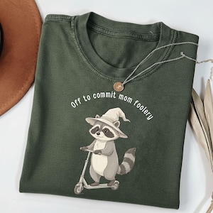 May include: Dark green t-shirt featuring a cartoon raccoon wearing a witch hat and riding a scooter. The text "Off to commit mom foolery" is arched above the raccoon. A silver necklace with a pendant is draped on the shirt.