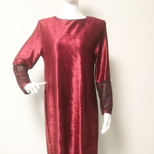 May include: A long, red velvet dress with black embroidered trim around the bottom hem and on the sleeves. The dress has a round neckline and long sleeves.
