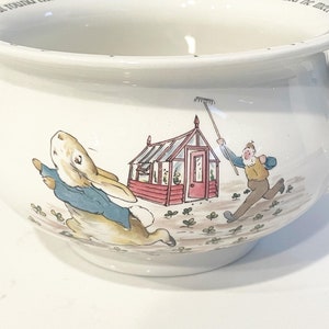 Può includere: Un vaso da fiori in ceramica bianca con un design di Peter Rabbit. Il vaso presenta un coniglio che scappa da un uomo con un rastrello, una serra rossa e trifoglio verde.