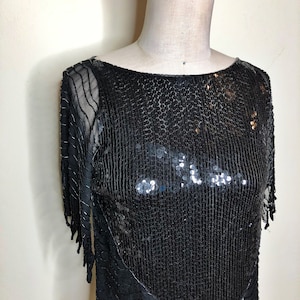 May include: A black sequined top with a scoop neck and short sleeves. The top has a fringe detail on the sleeves and hem.