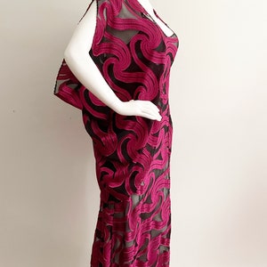 May include: A long, fuchsia-colored dress with a black sheer overlay. The dress has a swirling, abstract pattern and a loose, flowing silhouette.