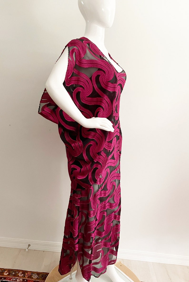 May include: A long, fuchsia-colored dress with a black sheer overlay. The dress has a swirling, abstract pattern and a loose, flowing silhouette.