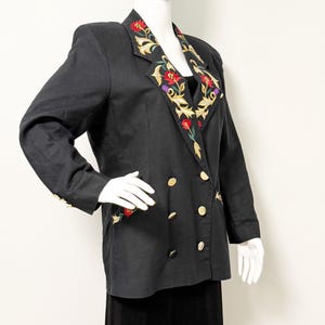 Puede incluir: Blazer negro con botones dorados y bordados florales en la solapa y los pu&ntilde;os. La chaqueta cruzada presenta un dise&ntilde;o bordado colorido con flores rojas, amarillas y moradas. El blazer se exhibe en un maniqu&iacute;.