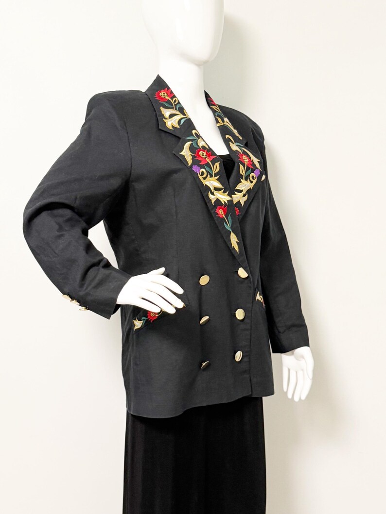 Puede incluir: Blazer negro con botones dorados y bordados florales en la solapa y los pu&ntilde;os. La chaqueta cruzada presenta un dise&ntilde;o bordado colorido con flores rojas, amarillas y moradas. El blazer se exhibe en un maniqu&iacute;.