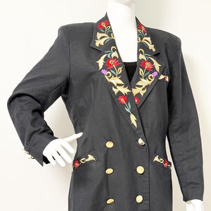 Puede incluir: Un blazer negro de doble botonadura con botones dorados y bordados florales en las solapas y los bolsillos. El bordado presenta flores rojas, doradas y moradas. El blazer se exhibe en un maniqu&iacute;.