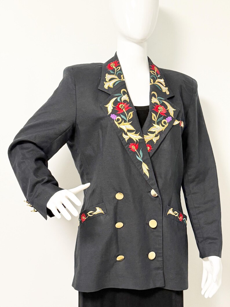 Puede incluir: Un blazer negro de doble botonadura con botones dorados y bordados florales en las solapas y los bolsillos. El bordado presenta flores rojas, doradas y moradas. El blazer se exhibe en un maniqu&iacute;.