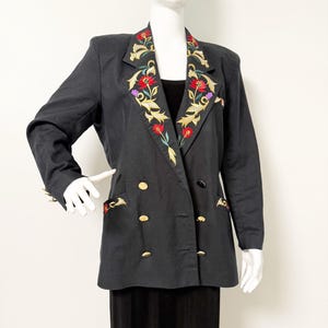 Puede incluir: Blazer negro con botones dorados y bordados florales en las solapas y bolsillos. El bordado presenta flores rojas, doradas y moradas con hojas verdes. El blazer es de doble botonadura y tiene un dise&ntilde;o cl&aacute;sico.