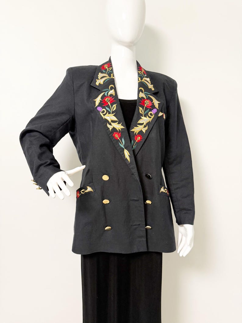 Puede incluir: Blazer negro con botones dorados y bordados florales en las solapas y bolsillos. El bordado presenta flores rojas, doradas y moradas con hojas verdes. El blazer es de doble botonadura y tiene un dise&ntilde;o cl&aacute;sico.