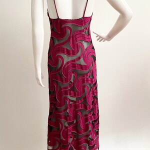 May include: A long, burgundy slip dress with a black sheer overlay featuring a swirling pattern. The dress has thin straps and a low back.