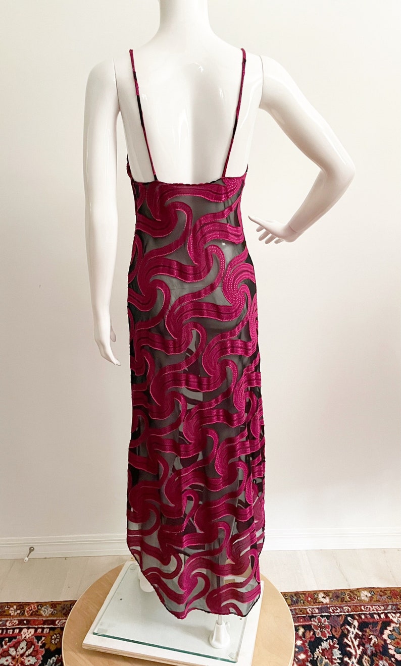 May include: A long, burgundy slip dress with a black sheer overlay featuring a swirling pattern. The dress has thin straps and a low back.
