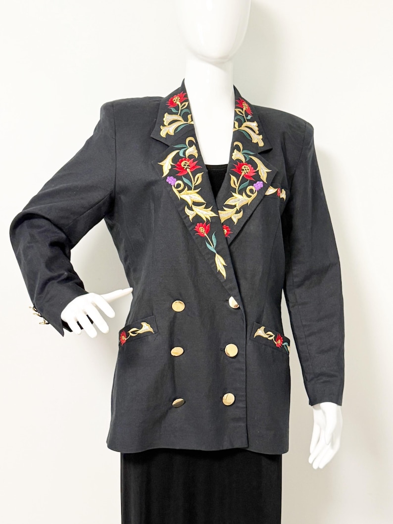 Puede incluir: Blazer negro de doble botonadura con botones dorados y bordados florales en las solapas y bolsillos. El bordado presenta colores rojo, dorado y verde. El blazer se exhibe en un maniqu&iacute;.