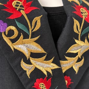 Puede incluir: Primer plano de un cuello de chaqueta negro con intrincado bordado floral. El bordado presenta flores rojas, hojas doradas y plateadas, y detalles morados. El dise&ntilde;o de la chaqueta es detallado y muestra una mezcla de colores y texturas.