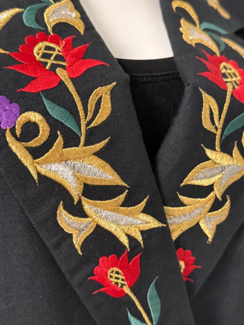 Puede incluir: Primer plano de un cuello de chaqueta negro con intrincado bordado floral. El bordado presenta flores rojas, hojas doradas y plateadas, y detalles morados. El dise&ntilde;o de la chaqueta es detallado y muestra una mezcla de colores y texturas.