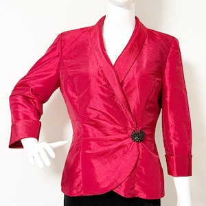 May include: A vibrant red blazer with a wrap-style front and three-quarter sleeves. The jacket features a decorative black embellishment at the waist and is displayed on a mannequin. The blazer is made of a shiny, textured fabric.