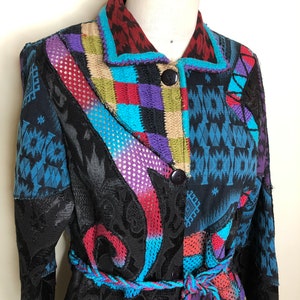 Puede incluir: Chaqueta patchwork colorida con un diseño único. La chaqueta presenta una mezcla de patrones y texturas, incluyendo formas geométricas, motivos florales y un cinturón trenzado. La paleta de colores incluye negro, azul, rojo, morado y beige.