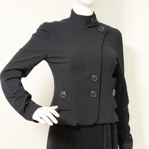 以下が含まれることがあります： ハイネックとボタン留めの黒いジャケット。 ジャケットは仕立ての良いフィット感で、構造的なデザインと小さなポケットが付いています。 ボタンは黒くて丸いです。 ジャケットはマネキンに展示されています。