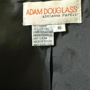 Puede incluir: Primer plano del interior de una prenda negra, con una etiqueta con la marca "ADAM DOUGLASS" y "ADRIANNA PAPELL". La etiqueta detalla la composici&oacute;n de la tela: 55% Ramio, 45% Algod&oacute;n y un forro 100% Acetato. La prenda es talla 10.