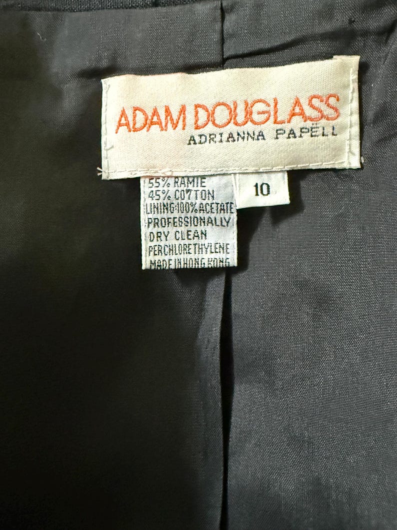 Puede incluir: Primer plano del interior de una prenda negra, con una etiqueta con la marca "ADAM DOUGLASS" y "ADRIANNA PAPELL". La etiqueta detalla la composici&oacute;n de la tela: 55% Ramio, 45% Algod&oacute;n y un forro 100% Acetato. La prenda es talla 10.