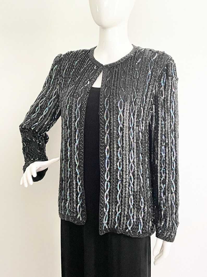 May include: A black sequined jacket with a subtle iridescent sheen. The jacket has a round neckline and long sleeves. The sequins are arranged in a pattern of vertical lines.