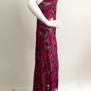 May include: A long, burgundy slip dress with a black sheer overlay featuring a swirling pattern. The dress has a thin strap and a fitted silhouette.