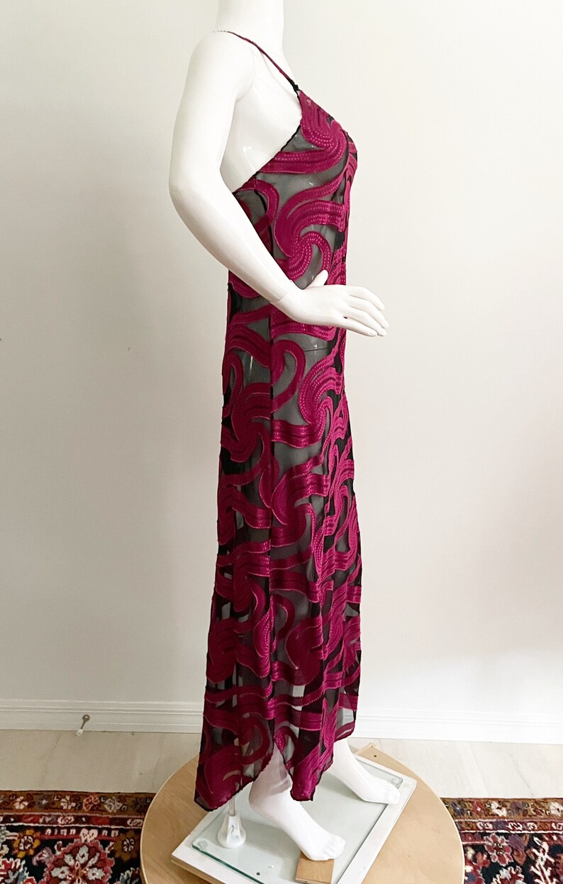 May include: A long, burgundy slip dress with a black sheer overlay featuring a swirling pattern. The dress has a thin strap and a fitted silhouette.
