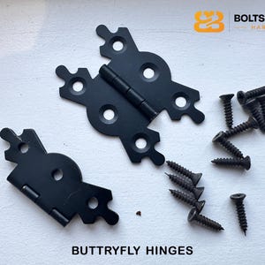 Op de afbeelding: Zwarte vlinderscharnieren en schroeven op een witte ondergrond. De scharnieren hebben de vorm van een vlinder, met meerdere schroefgaten. De tekst "BUTTRYFLY HINGES" staat onderaan. Het logo van Bolts & Brass Hardware staat rechtsboven.