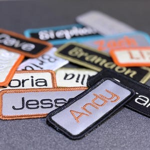 May include: A collection of rectangular embroidered name patches in various colors including black, white, grey, and blue. Each patch features a stitched name such as Dave, Victoria, Jess, Andy, Sarah, Brandon, Liz, and Zach in contrasting thread.