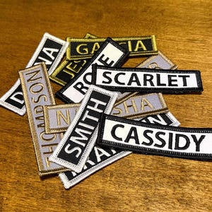 May include: A collection of rectangular embroidered name patches featuring various names including SCARLET, SMITH, and CASSIDY. The patches have black or white backgrounds with contrasting text and stitched borders in gold, white, or black thread.