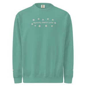 May include: A teal sweatshirt with the phrase "repulsively cheerful woman" embroidered in white, surrounded by small daisy-like flowers. The long-sleeved top has a crew neck and ribbed cuffs.