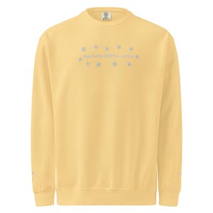 May include: A light yellow sweatshirt with the text "reputedly cheerful woman" in light blue, surrounded by snowflake designs. The long-sleeved crew neck shirt is a solid color.