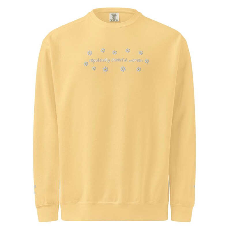 May include: A light yellow sweatshirt with the text "reputedly cheerful woman" in light blue, surrounded by snowflake designs. The long-sleeved crew neck shirt is a solid color.