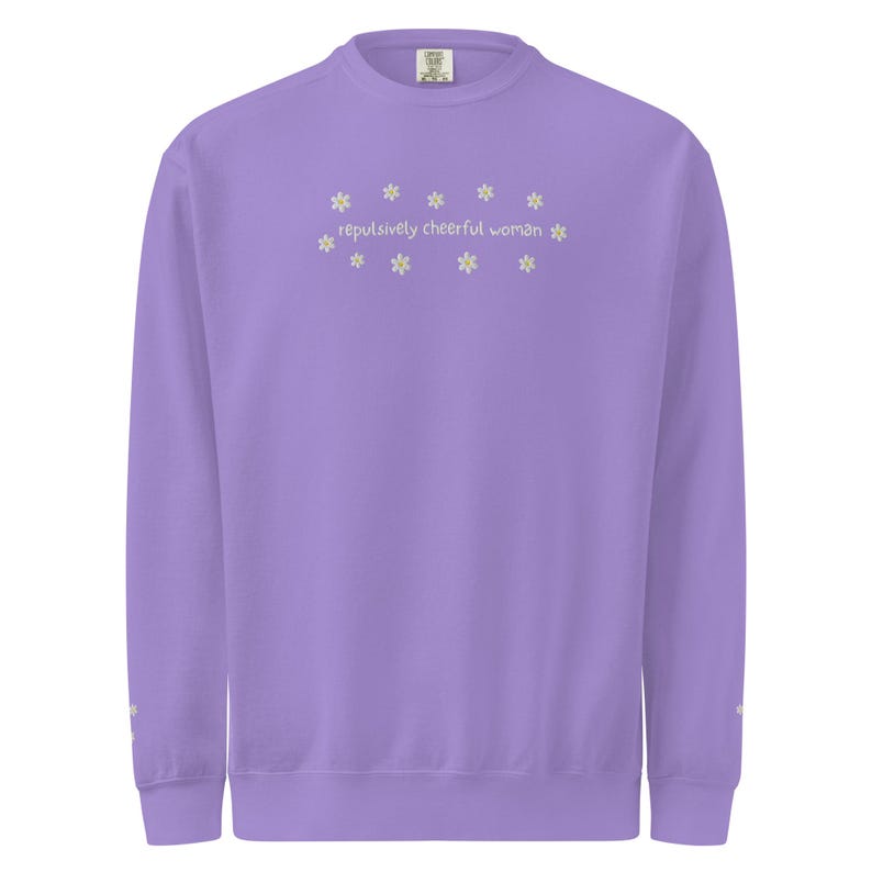 May include: A lavender sweatshirt with the text "repulsively cheerful woman" embroidered in a light yellow color, surrounded by small daisy flower designs. The sweatshirt has a crew neck and long sleeves.