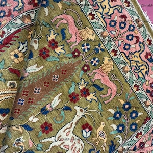 May include: Close-up of a hand-woven rug with intricate floral and animal designs. The rug features a central olive green field with pink, red, blue, and yellow accents. The border is a mix of pink and green, with a fringed edge.