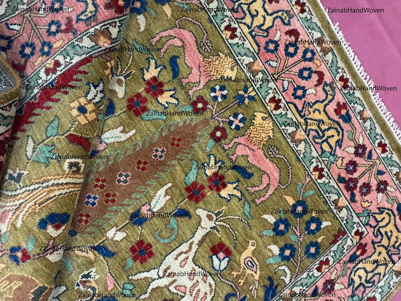 May include: Close-up of a hand-woven rug with intricate floral and animal designs. The rug features a central olive green field with pink, red, blue, and yellow accents. The border is a mix of pink and green, with a fringed edge.