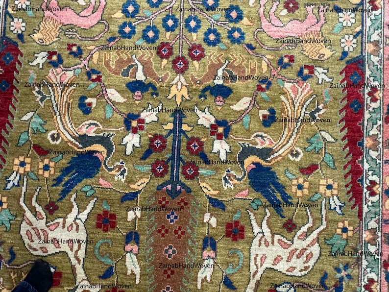 May include: Close-up of a hand-woven rug with an intricate design. The rug features a central tree-like motif with blue, red, and yellow floral elements. The background is olive green, with animal and bird figures.