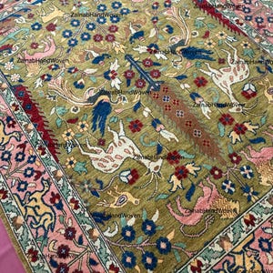 May include: An olive green rug with a detailed pattern featuring floral and animal motifs in shades of pink, blue, and red. The rug has a border with a repeating floral design. The text "ZainabHandWoven" is visible in the image.
