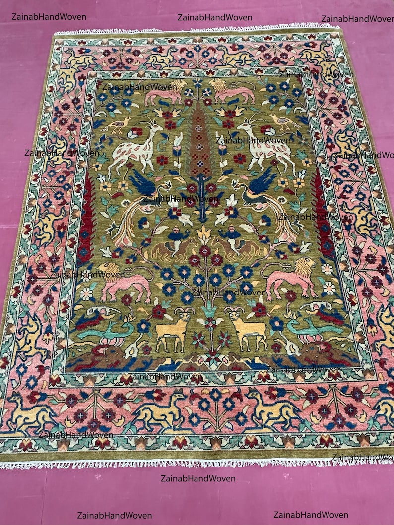 May include: Hand-woven rug with a central olive green field featuring stylized animal and floral motifs in shades of pink, blue, yellow, and brown. The border is a complementary pink with repeating floral patterns. The rug has a fringed edge.