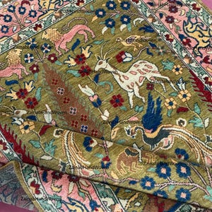 Puede incluir: Una alfombra tejida a mano en verde oliva con un detallado patrón floral y animal. El diseño central presenta ciervos y pájaros, rodeados por un borde de motivos florales rosas, azules y amarillos. Las medidas no son visibles.